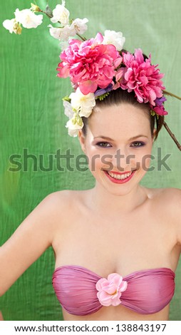 Beauty portrait of a retro young woman wearing a spring flowers hair dress while wearing a flower top, standing against a green fabric background, smiling.