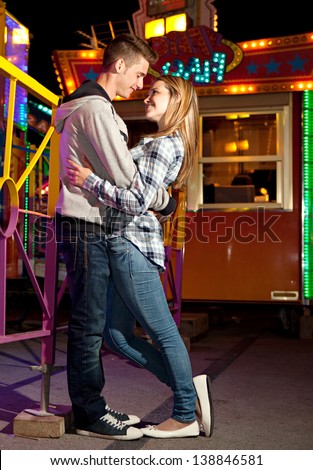Young couple visiting a funfair ground arcade being romantic and hugging with rides and lights in the background, during the evening night time.