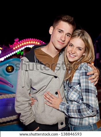 Portrait of a young teenager couple visiting an attractions park arcade and getting excited while standing next to a colorful ride at night time.