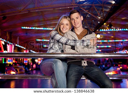 Young couple sitting by a bouncy cars ride while visiting an attractions park ground with their heads together and smiling with colorful lights in the background at night time.