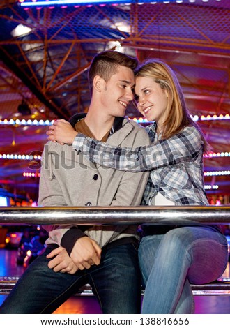 Young couple sitting by a bouncy cars ride while visiting an attractions park ground with their heads together and smiling with colorful lights in the background at night time.
