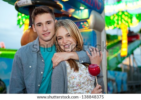 Close up portrait of a young couple visiting a funfair ground arcade and hugging while holding a sugar red apple sweet with rides and lights in the background, during evening night time.