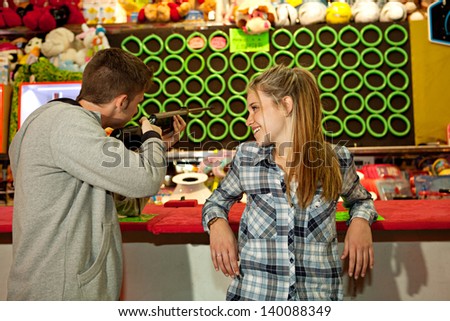 Young couple playing shooting games while visiting an amusement park arcade at night time together, having fun and competing, with prizes in the background