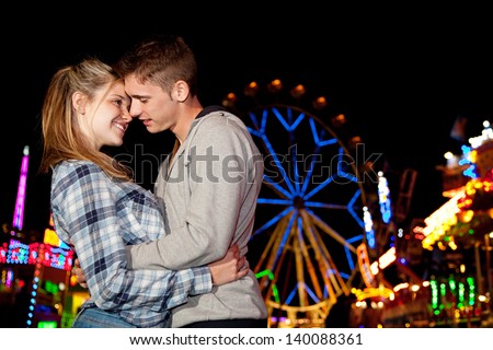 Young couple hugging while visiting an attractions park arcade at night time, with colorful lights and rides around them.
