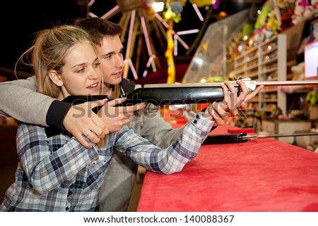 Young couple playing shooting games while visiting an amusement park arcade at night time, having fun with color lights and rides in the background, man helping woman.