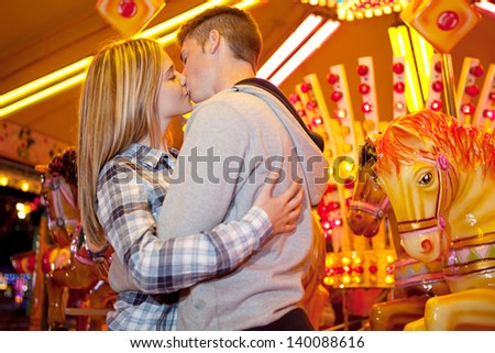Young couple visiting an amusement park arcade together, hugging and kissing while standing next to a carousel ride with lights at night.