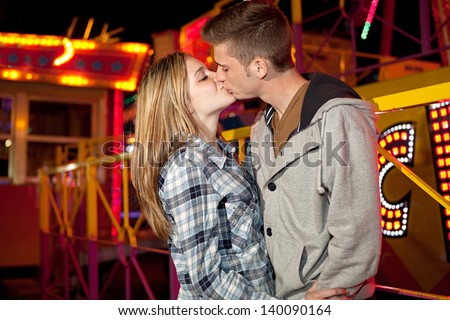 Portrait of a young couple visiting an attractions park arcade being romantic and kissing each other with rides and lights in the background during night time.