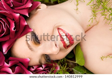 Close up beauty portrait of a young beautiful woman laying down in a forest wearing a red roses head dress hat while relaxing on a green bed of leaves and grass, joyful and smiling.