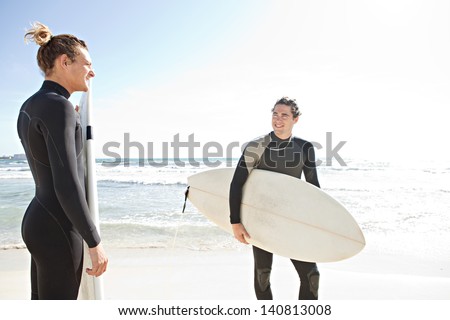 Two surfers friends sharing a surfing trip experience, holding their surfing boards on a sunny white sand beach with an intense blue sky while on vacation and wearing specialist black neoprene suits.