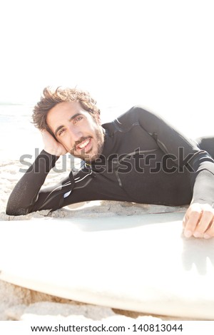 Young surfer sport man laying down and relaxing next to his surfing board on a white sand beach during a sunny day, smiling at camera.