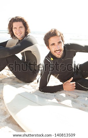 Two joyful surfers friends sharing a surfing trip experience, laying down next to their surfing boards on a beach with a sunny sky while on vacation and wearing specialist black neoprene suits.