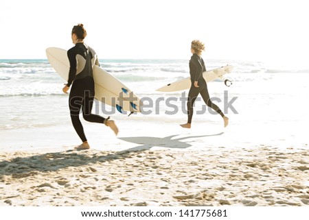 Two young surfers running together towards the ocean while carrying their surfing boards under their arms during a sunny day on a white sand beach.