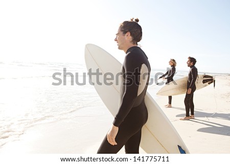 Three sport surfer friends standing together on a white sand beach carrying their surfing boards near the shore during a sunny golden day on vacation, looking out at sea.