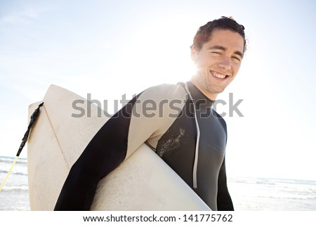 Young surfer man carrying his surfing board under his arm and walking away from the sea during a sunny day with a blue sky on vacation.