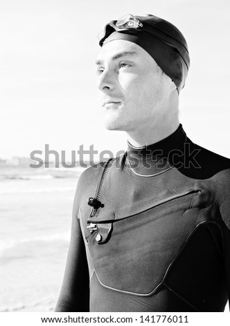 Black and white portrait of professional swimmer wearing a neoprene suit and a rubber hat getting ready for training, standing on a white sand beach during a sunny day.