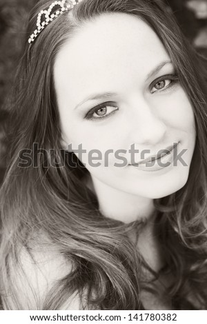 Black and white close up beauty portrait of a young attractive woman wearing cosmetics and a diamond tiara, looking at camera and smiling with lipstick.