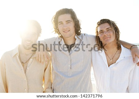 Three young men friends hanging around on vacation together with their arms around each other at sunset while on a beach having fun and smiling during the summer.