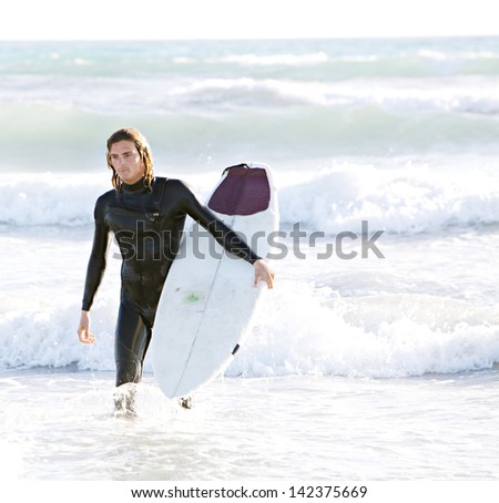 Attractive surfer walking back to shore after a surfing session in the sea with crashing waves around him, feeling powerful and focused.