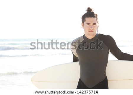 Portrait of a young attractive surfer man standing on a beach and carrying his surfing board while looking at camera during a sunny day on vacation.