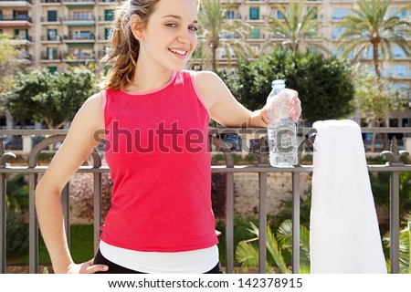 Happy young woman taking a break from exercising in the city, holding a blue bottle of mineral water and smiling at the camera during a sunny day.