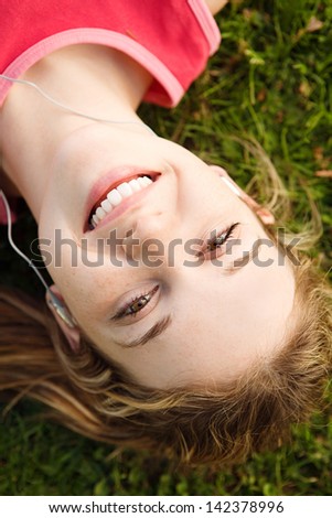 Close up over head view of a joyful young woman laying down on a park green grass, taking a break from exercising while listening to music with her mp4 player and earphones.