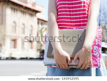 Close up middle section view of a teenager student carrying homework, books and note pads while standing near a college university building in a campus during a sunny summer day.