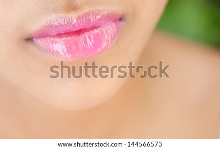 Close up detail view of a beautiful young woman's mouth with perfect lips wearing glossy pink lipstick against a nature green background.