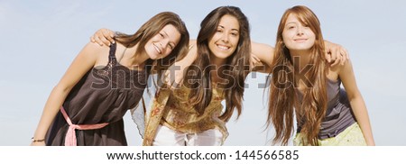 Panoramic wide format portrait of a group of three girls friends smiling to the camera, standing together against a blue sky with their arms around each others shoulders during their summer vacation.