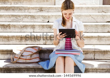 Portrait of a teenager student girl using with her digital tablet to go on-line while sitting down on a university campus entrance stone steps, smiling during  a sunny day.