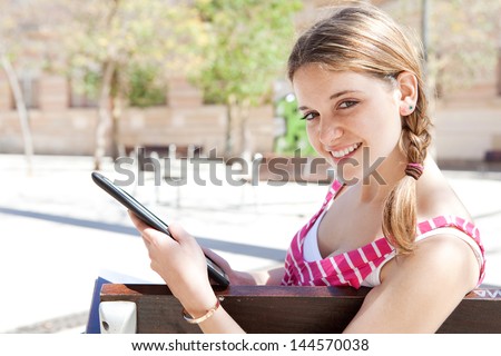 Side portrait of a teenager student using a digital tablet pad to go on-line while sitting on a wooden bench in a campus exterior during a sunny day, smiling.