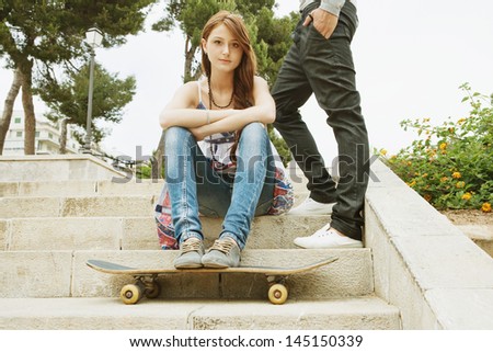 Two young fashionable teenager friends relaxing in an urban city park with their skating board, lounging and socializing during a summer weekend, girl being thoughtful.