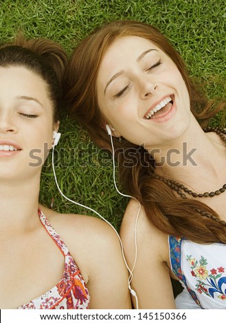 Two teenager girls relaxing with their heads together laying down on green grass in a park, sharing their headphones and singing to the music, having fun and enjoying the song.