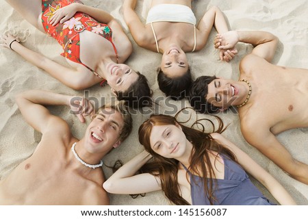 Over head view of a group of five mixed teenagers laying down on a fine white sand beach with their heads together, smiling at the camera during their summer vacation away.