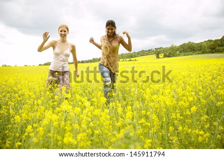 Two teenager girls friends running together on a large yellow flowers field during a summer vacation in the countryside on a sunny day, having fun.