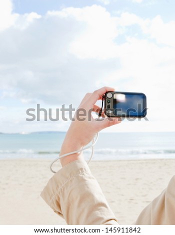Young woman land holding a consumer digital photo camera with a blank screen and pointing at the sea while visiting a white sand beach on vacation, rear view.