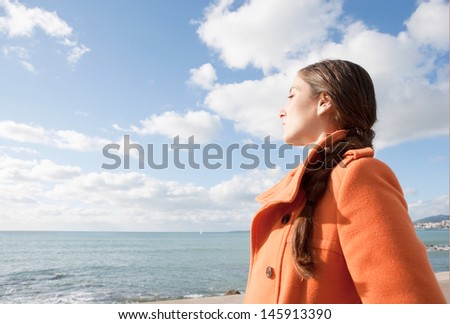 Profile portrait view of a young woman facing the sea and breathing fresh air with her eyes closed against an intense blue sky during a sunny winter day and wearing an orange coat.