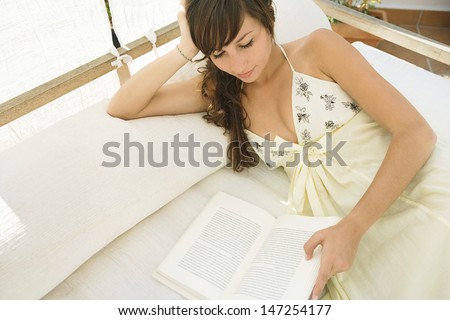 Attractive young woman relaxing on an outdoors bed in the shade, reading a book and relaxing during her summer vacation.