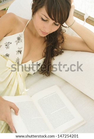 Attractive young woman relaxing on an outdoors bed in the shade, reading a book and relaxing during her summer vacation.