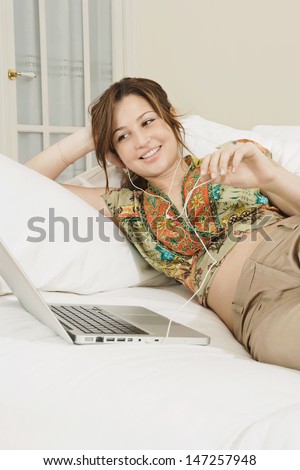 Beautiful young woman lounging on her sofa at home and listening to music with her laptop computer and headphones, smiling and enjoying the moment.
