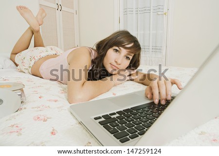 Young beautiful teenager woman laying down on her bed at home using a pc laptop computer to listen to music while relaxing in her bedroom.
