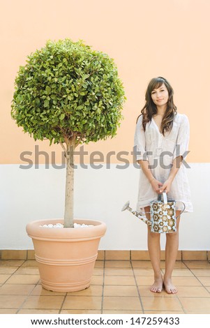 Attractive young woman standing proudly next to her topiary tree plant on a roof terrace holding a full watering can while doing her gardening, smiling.