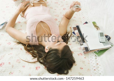 Over head view of a beautiful teenager relaxing and laying down on her bed in her bedroom at home, using an mp3 player and headphones to listen to music.