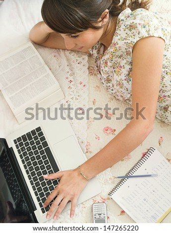 Over head view of a teenager student doing her homework, laying on her bed in her bedroom, using a laptop computer and having her books and mobile phone near her.