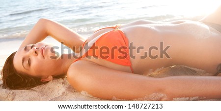 Horizontal format profile view of a sexy young woman laying down and relaxing on a beach while on vacation, wearing an orange bikini and sunbathing with the sunset light, exterior.