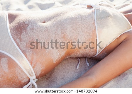 Middle section of a woman body covered in white fine sand while laying down on a beach wearing a luxury sequins bikini and looking sexy, relaxing on vacation, faceless view.