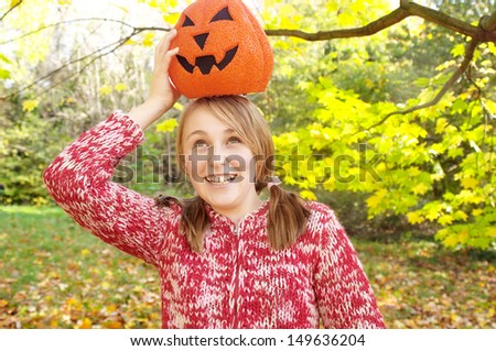Joyful girl holding a fake halloween pumpkin on top of her head and having fun while visiting a forest park with falling leaves and autumn colors during a sunny holiday day.