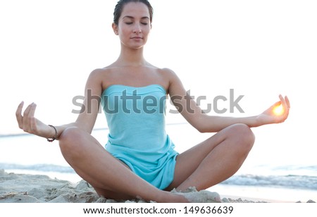Low angle view of a healthy young woman sitting in a yoga position meditating, with the sun filtering through her fingers in a circle during sunset on a tranquil beach.
