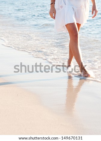 Lower body section of a young woman walking along a white sand beach shore at sunset, taking steps and relaxing in an idyllic destination during a summer vacation.