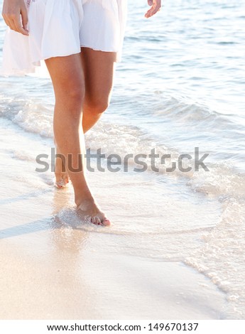 Lower body section of a young woman walking along a white sand beach shore at sunset, taking steps and relaxing in an idyllic destination during a summer vacation.