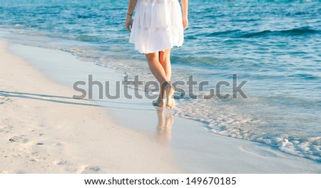 Rear lower body section of a young woman walking along a white sand beach shore at sunset, taking steps and relaxing with an intense blue sea during a summer vacation.